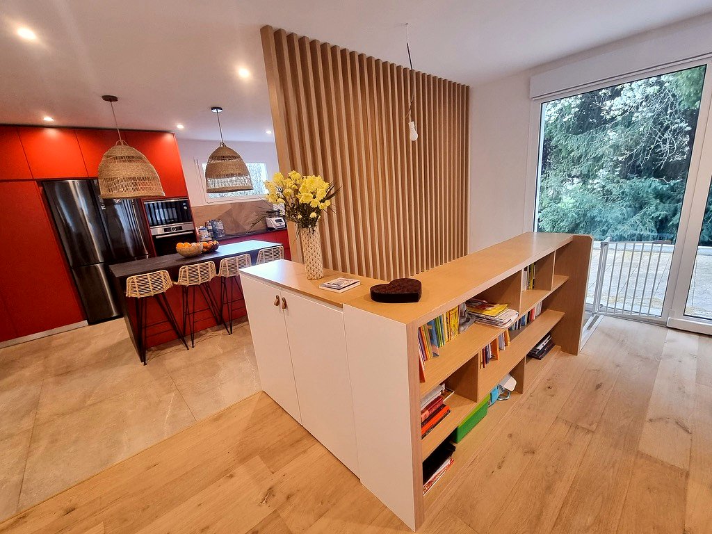Séjour ouvert sur la cuisine rouge avec îlot, séparé par un claustra bois et un meuble bibliothèque, parquet chêne.